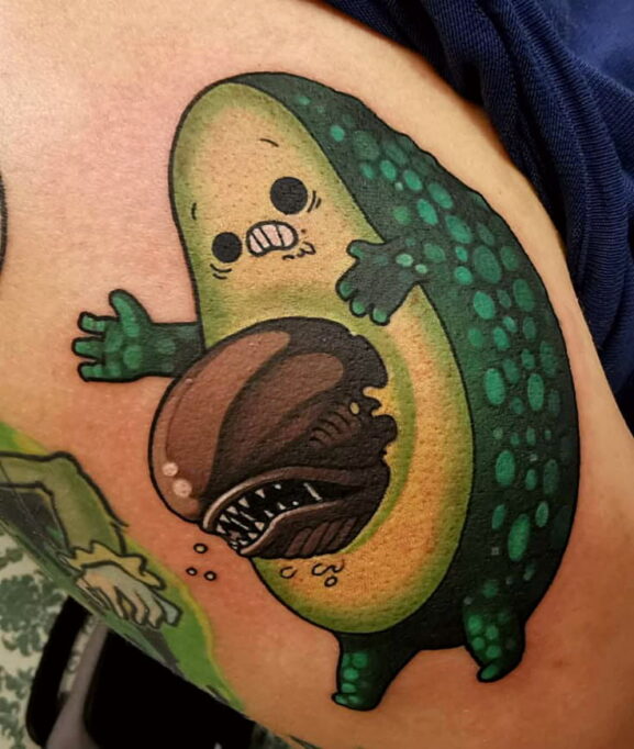 Avocado Character: The main figure is a cartoon-style anthropomorphic avocado, complete with arms, legs, and an expressive face. Its facial features—round eyes and clenched teeth—convey a look of fear or surprise. The texture on the avocado skin is exaggerated with large, teal-green spots for a playful, stylized effect. Chest-Bursting Alien Element: Emerging from the avocado’s seed area is a menacing creature resembling the Xenomorph chestburster from Alien. The creature has a shiny, dark brown tone with sharp teeth and a ridged head, appearing to erupt from inside the avocado. Droplets and shading add a slimy, visceral quality to the effect. Art Style: The tattoo uses bold outlines, vibrant saturation, and clean shading, aligning with New School tattoo aesthetics—a style known for its cartoonish proportions and exaggerated themes. The linework is crisp, and the colors are bright, contributing to the comic-book-like feel. Placement: The tattoo appears to be on the thigh or upper leg area, providing ample space for detail and expressive composition. Theme & Interpretation: This tattoo mixes cuteness with horror, reflecting a love for pop culture parody, especially dark sci-fi and alien-themed stories. It could symbolize: A playful take on internal struggle or surprise Love for dark humor and unexpected contrasts A celebration of geek culture, particularly fandoms like Alien